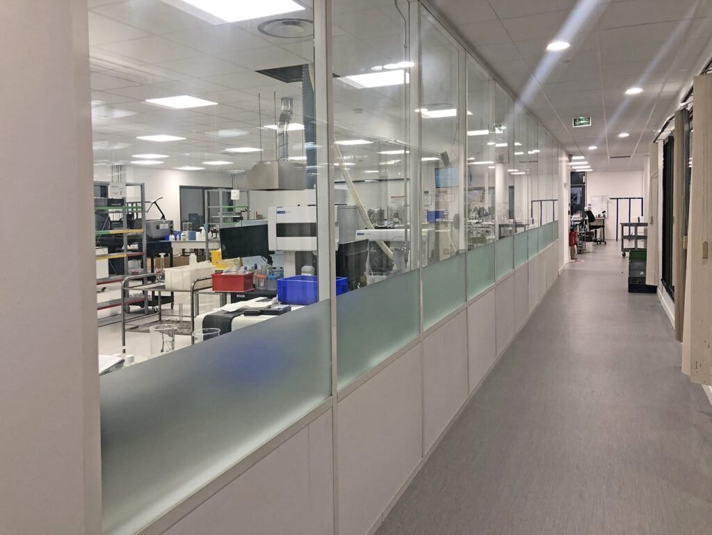 Un laboratoire moderne avec de grandes baies vitrées, des postes de travail visibles, des équipements et des conteneurs de stockage ; un couloir lumineux longe le laboratoire.