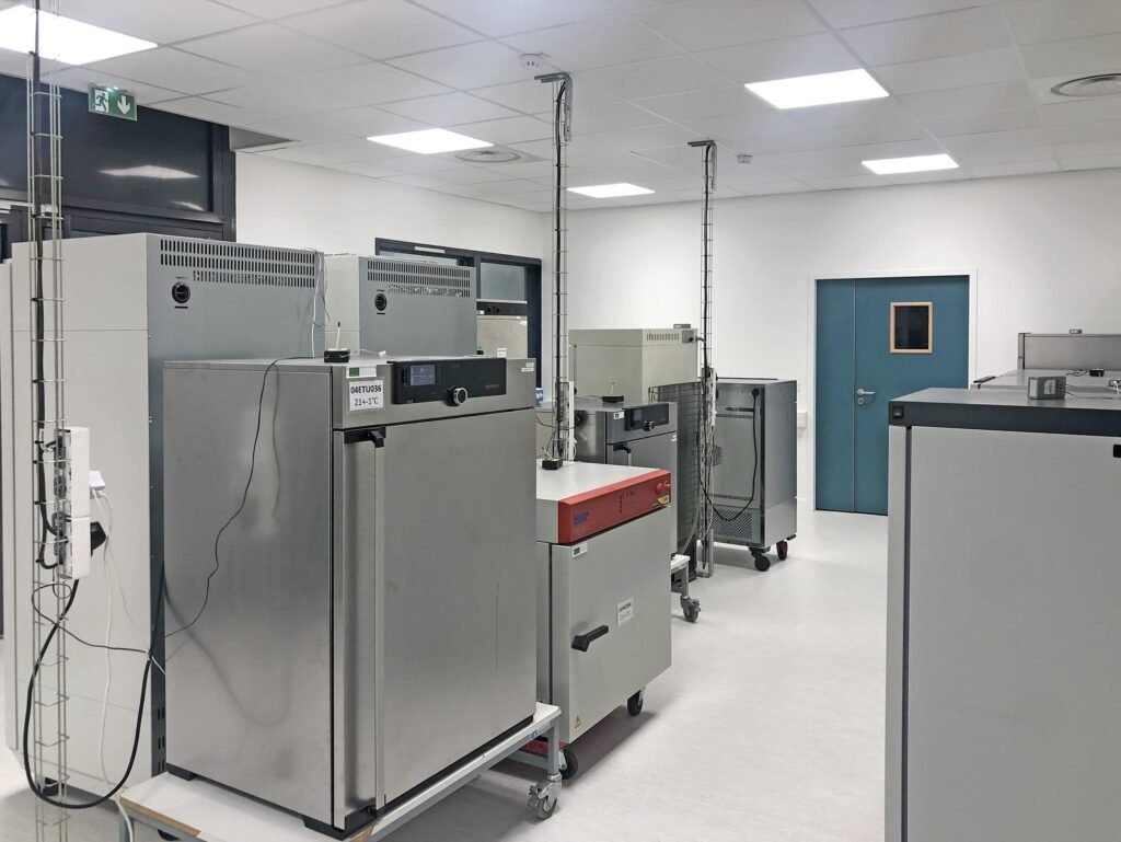 Une salle de laboratoire avec plusieurs grands fours de laboratoire en acier inoxydable, des incubateurs et des équipements sur roues, sous des plafonniers lumineux.