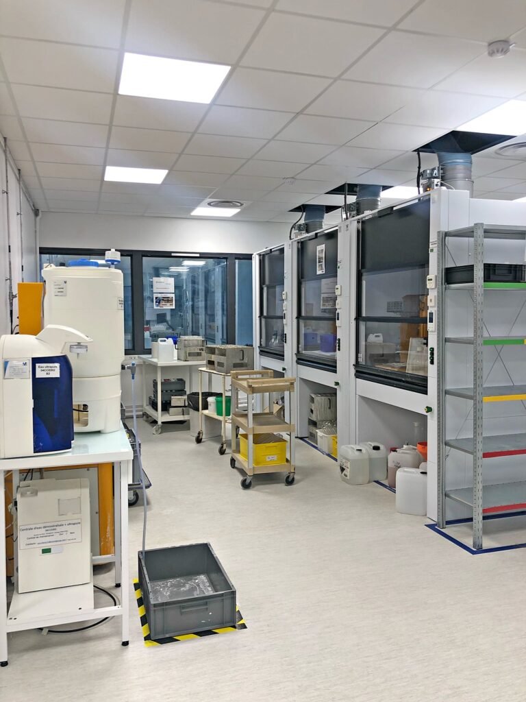 Un laboratoire propre et organisé avec des postes de travail, des étagères, du matériel et des conteneurs de stockage de sécurité sous un éclairage zénithal lumineux.