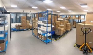 Une salle d'entrepôt avec des étagères et des palettes contenant des cartons empilés, du matériel d'emballage et un transpalette jaune sur le sol.