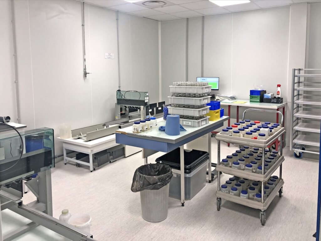 Une salle de laboratoire avec des tables de travail, des plateaux contenant des échantillons, du matériel de laboratoire, un écran d'ordinateur, des supports métalliques et une poubelle.