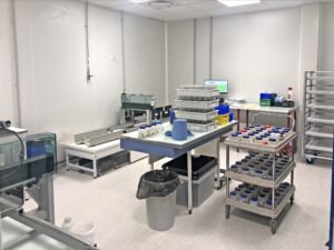 Une salle de laboratoire avec des tables de travail, des plateaux contenant des échantillons, du matériel de laboratoire, un écran d'ordinateur, des supports métalliques et une poubelle.