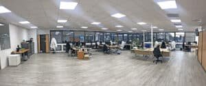 Bureau spacieux, moderne et ouvert, avec des bureaux, des chaises, des ordinateurs, de grandes fenêtres et un éclairage zénithal puissant ; personne n'est présent.