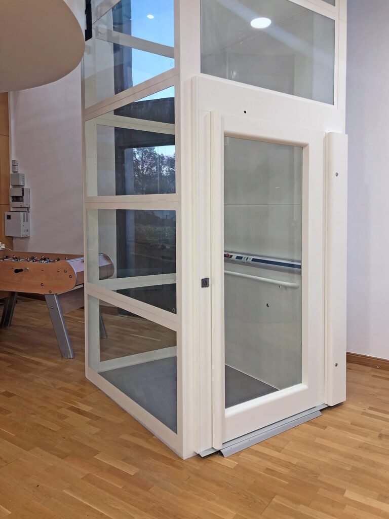 Un ascenseur domestique moderne en verre avec un cadre blanc est installé à l'intérieur sur un sol en bois, à côté d'un baby-foot.