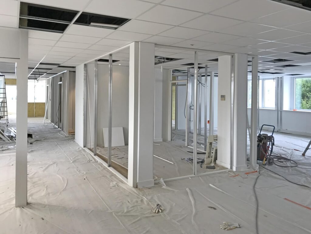 Espace de bureaux en cours de rénovation avec des dalles de plafond apparentes, des murs inachevés et des matériaux de construction éparpillés sur les sols couverts.