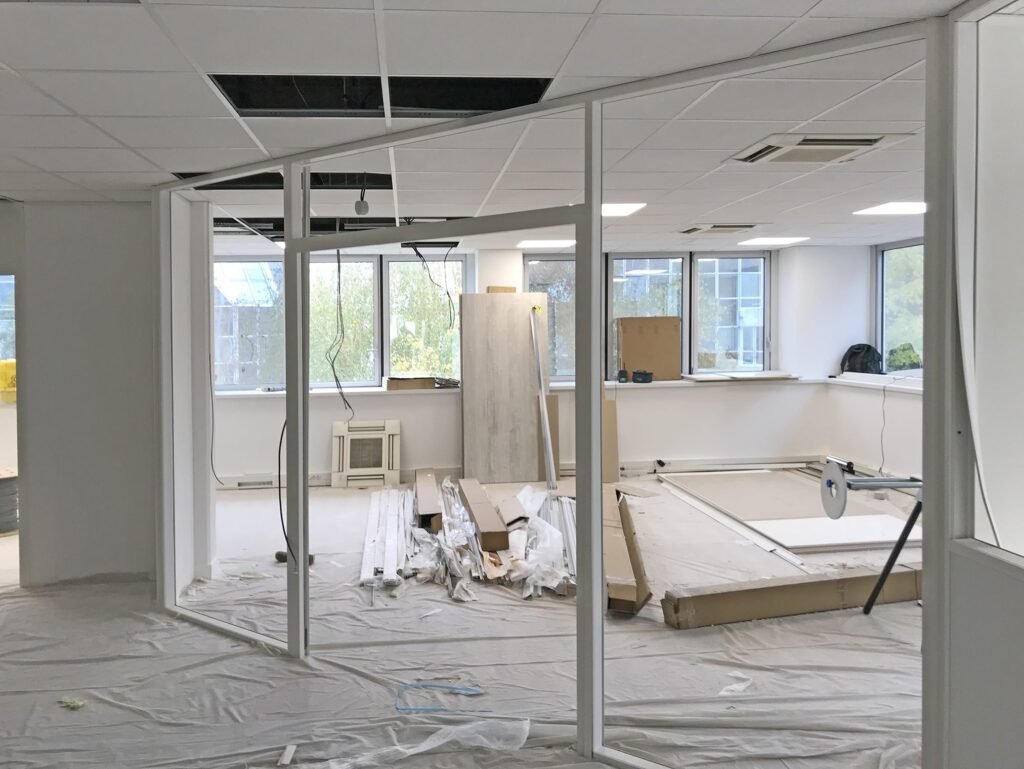 Espace de bureaux en cours de rénovation avec des dalles de plafond apparentes, des matériaux de construction et des outils éparpillés sur le sol près de grandes fenêtres.