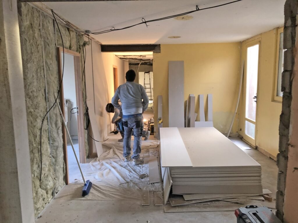 Deux personnes travaillant dans une pièce en cours de rénovation, avec de l'isolant sur un mur, des plaques de placoplâtre empilées sur le sol et des outils et matériaux de construction éparpillés.