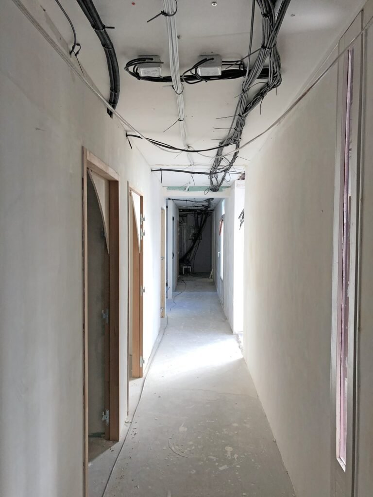 Couloir non aménagé avec des câbles apparents au plafond, des murs non peints et des portes menant à des pièces en construction.