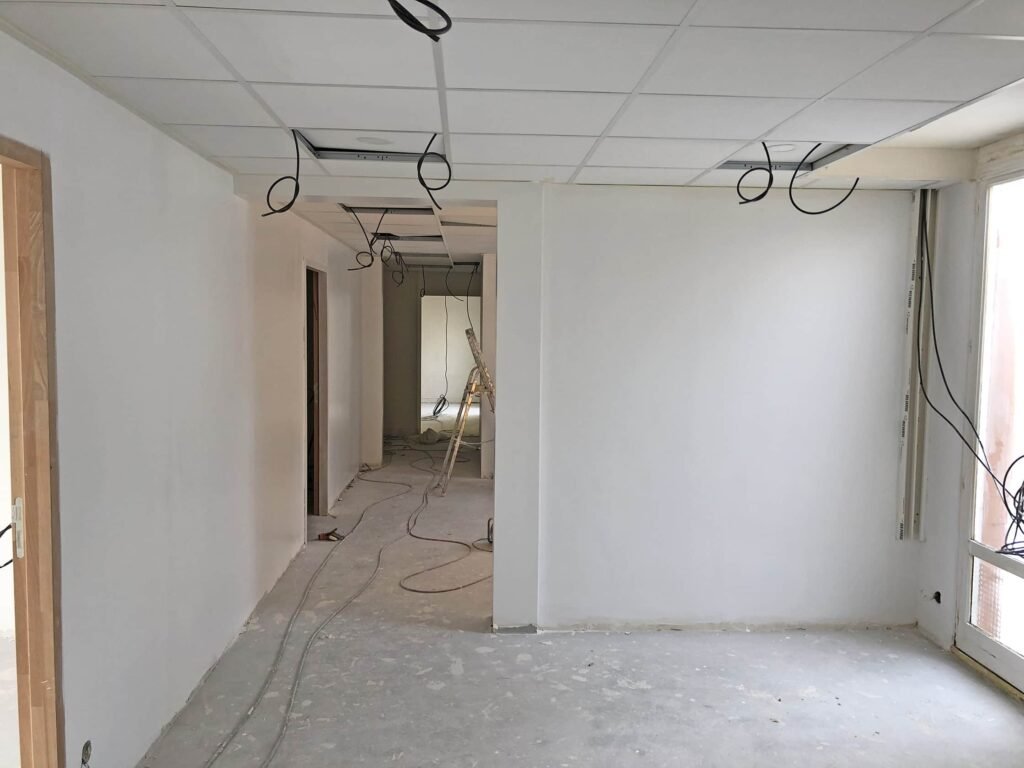 Une pièce en cours de rénovation avec des murs blancs, des sols non finis, des fils électriques apparents qui pendent du plafond et des matériaux de construction visibles à l'arrière-plan.