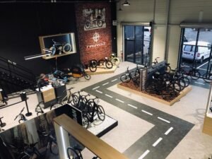 Une salle d'exposition présente divers vélos, y compris des vélos électriques, avec une route peinte sur le sol, des accessoires pour vélos et des affiches promotionnelles sur les murs.