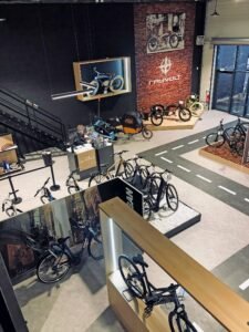 Intérieur d'un magasin de vélos moderne avec plusieurs vélos en exposition, une fausse piste au sol, des vélos fixés au mur et un comptoir de caisse près d'un escalier.