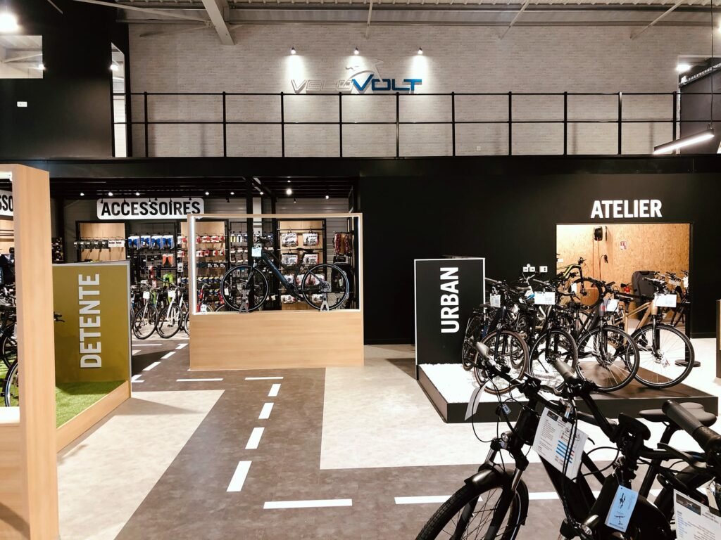 Intérieur d'un magasin de vélos moderne avec des sections intitulées Détente, Accessoires, Urban et Atelier. Plusieurs vélos et accessoires sont exposés dans le magasin.