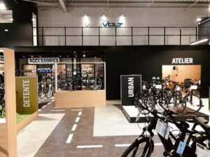 Intérieur d'un magasin de vélos moderne avec des sections intitulées Détente, Accessoires, Urban et Atelier. Plusieurs vélos et accessoires sont exposés dans le magasin.