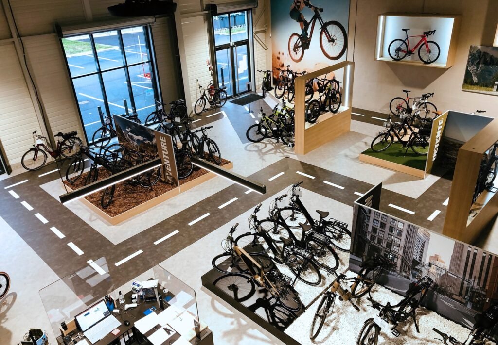 Vue large d'une salle d'exposition de bicyclettes moderne avec divers vélos exposés sur des supports, des marquages routiers intérieurs et de grandes affiches sur le cyclisme sur les murs.