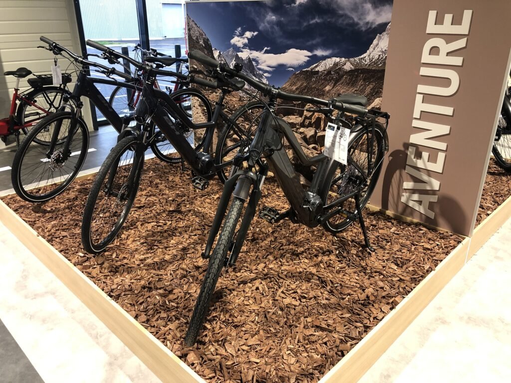 Vélos électriques présentés sur un sol en copeaux de bois dans une salle d'exposition, avec un grand panneau "AVENTURE" et un paysage de montagne en toile de fond.