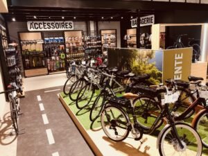 Intérieur d'un magasin de bicyclettes avec des rangées de vélos en exposition, une section d'accessoires et une enseigne de boulangerie visible à l'arrière-plan.