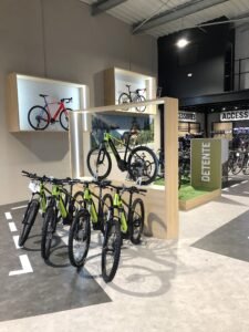 Exposition de vélos tout-terrain dans un magasin de vélos moderne, avec plusieurs vélos verts au premier plan et deux vélos montés sur des étagères en bois à l'arrière-plan.