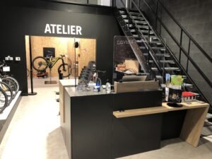 Intérieur d'un magasin de vélos moderne avec un comptoir de service, un écran de protection et un escalier ; un atelier avec des vélos est visible à l'arrière-plan sous un panneau portant l'inscription "ATELIER".
