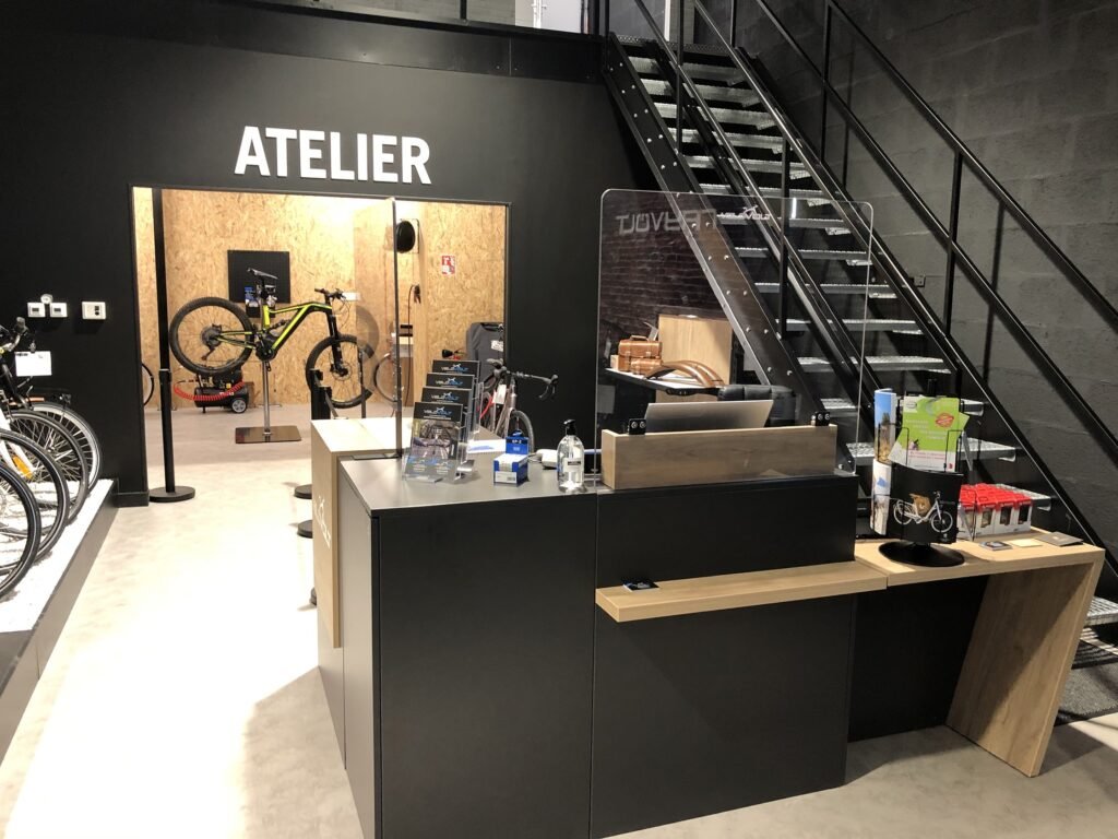 Intérieur d'un magasin de bicyclettes moderne avec une réception, un escalier et un atelier portant la mention "Atelier" à l'arrière-plan. Des bicyclettes et des outils de réparation sont visibles.