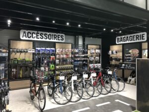 Un magasin de vélos avec plusieurs vélos devant et des étagères d'accessoires et de sacs étiquetés "Accessoires" et "Bagagerie" à l'arrière-plan.