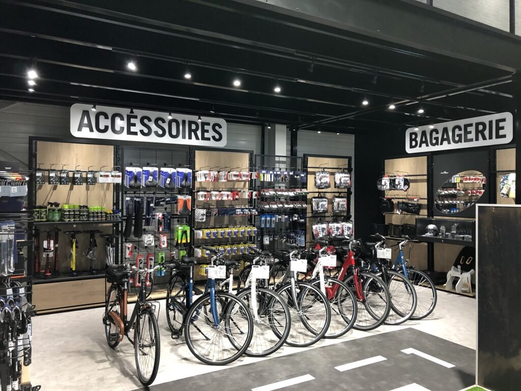 Un étalage de vente au détail avec des bicyclettes devant et des étagères d'accessoires de cyclisme et de sacs étiquetés "ACCESSOIRES" et "BAGAGERIE" dans un magasin bien éclairé.