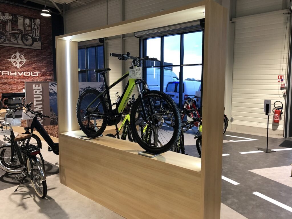 Un vélo tout-terrain à pneus épais est exposé sur un support en bois dans une salle d'exposition, avec d'autres vélos visibles à l'arrière-plan.