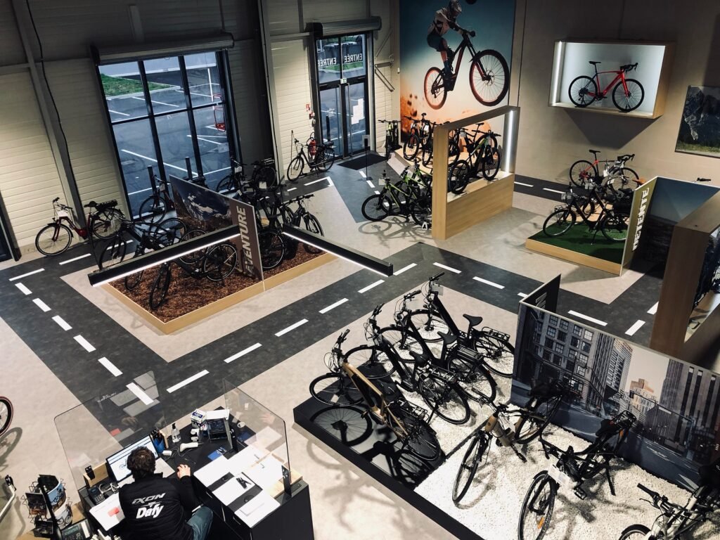 Intérieur d'un magasin de bicyclettes avec plusieurs bicyclettes exposées sur des supports et des étagères, une personne travaillant à un bureau et des décorations sur le thème du cyclisme sur les murs.