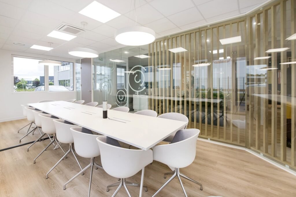 Salle de conférence moderne avec une longue table blanche, douze chaises blanches, des lampes suspendues, des murs en verre et un sol en bois. De grandes fenêtres apportent de la lumière naturelle.
