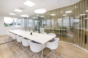 Salle de conférence moderne avec une longue table blanche, douze chaises blanches, des lampes suspendues, des murs en verre et un sol en bois. De grandes fenêtres apportent de la lumière naturelle.