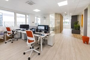 Espace de bureau moderne avec bureaux, ordinateurs et chaises orange ; murs blancs, grandes fenêtres, parquet en bois clair et plantes en pot.