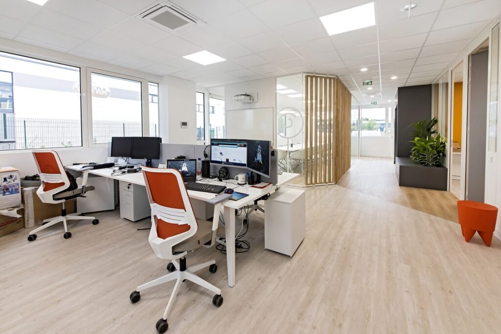Espace de bureau moderne avec bureaux, ordinateurs, chaises orange et blanches, grandes fenêtres, plantes et cloisons en verre.