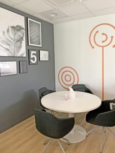 Un petit espace de réunion avec quatre chaises sombres autour d'une table ronde blanche, de l'art mural sur un mur gris, un sol en bois et des motifs circulaires orange sur un mur blanc.