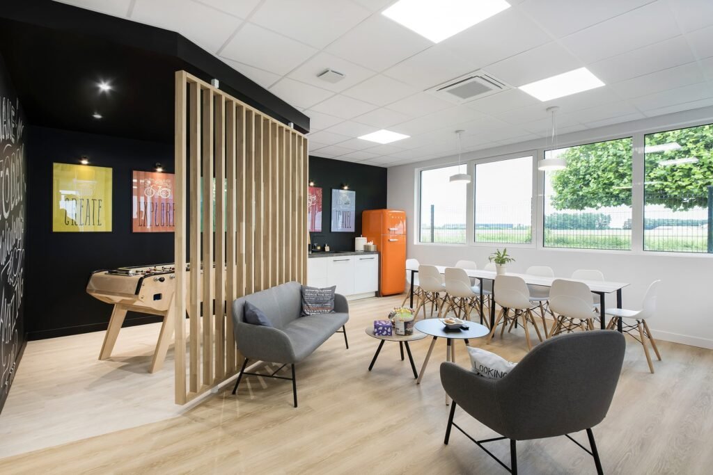 Une salle de pause avec un baby-foot, un coin salon, une cuisine avec un réfrigérateur rétro orange et une table à manger près de grandes fenêtres.