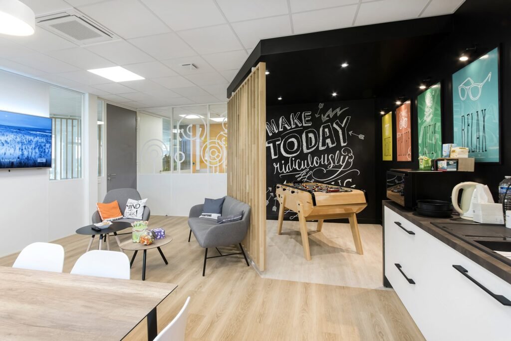 Salle de pause de bureau moderne avec des chaises longues, un baby-foot, une petite kitchenette, des œuvres d'art murales et des snacks sur une table, avec un sol en bois clair et un éclairage lumineux.
