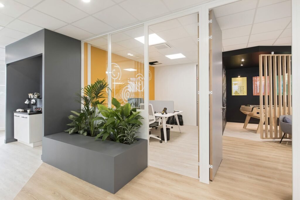 Bureau moderne avec des murs en verre, des plantes d'intérieur, des bureaux avec des chaises, un baby-foot et un sol en bois.