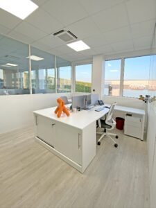 Bureau moderne avec un bureau blanc, des écrans d'ordinateur, une chaise de bureau, une figurine orange en forme de chien et de grandes fenêtres laissant entrer la lumière naturelle.