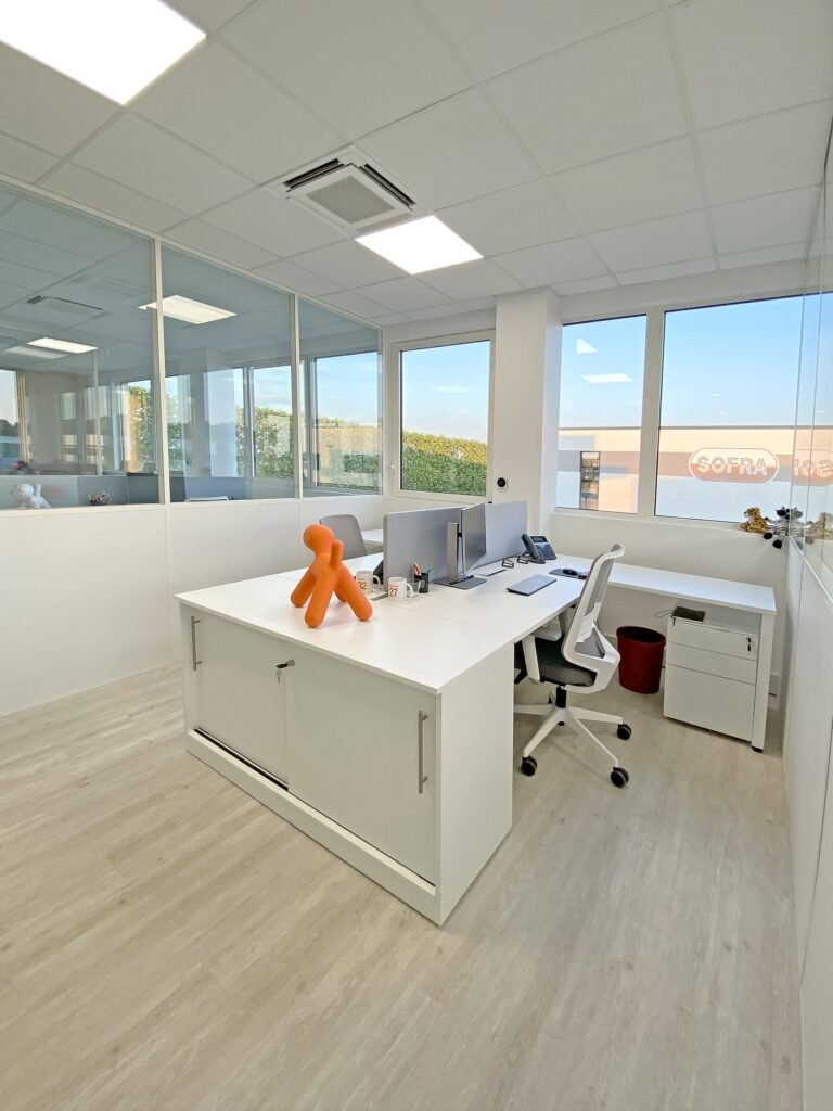 Bureau moderne avec un bureau blanc, des écrans d'ordinateur, une chaise de bureau, une figurine orange en forme de chien et de grandes fenêtres laissant entrer la lumière naturelle.