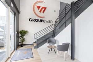 Entrée de bureau moderne avec deux chaises grises, une petite table ronde, une plante en pot et un escalier. Logo "GROUPE WIO" affiché sur un mur de briques blanches.