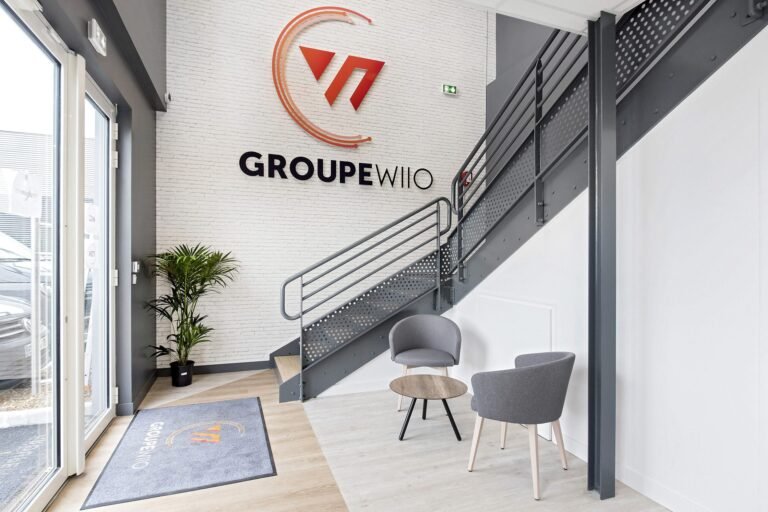 Entrée de bureau moderne avec deux chaises grises, une petite table ronde, une plante en pot et un escalier. Logo "GROUPE WIO" affiché sur un mur de briques blanches.