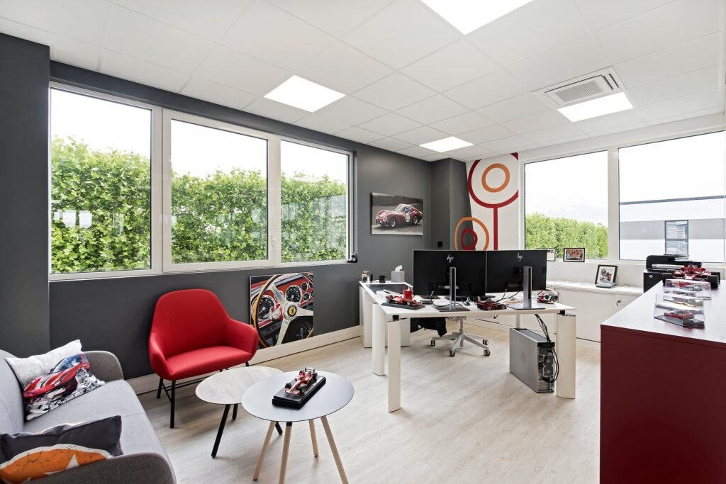 Bureau moderne avec de grandes fenêtres, deux bureaux avec des ordinateurs, un fauteuil rouge, un canapé gris et une décoration sur le thème de l'automobile sur les murs et les tables.