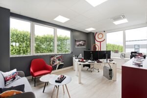 Bureau moderne avec de grandes fenêtres, deux bureaux avec des ordinateurs, un fauteuil rouge, un canapé gris et une décoration sur le thème de l'automobile sur les murs et les tables.