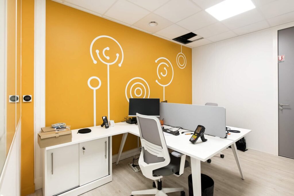 Bureau moderne avec deux bureaux blancs, des chaises grises, des écrans d'ordinateur et des téléphones. Mur d'accent jaune avec des motifs abstraits blancs ; panneau de plafond manquant au-dessus de la tête.