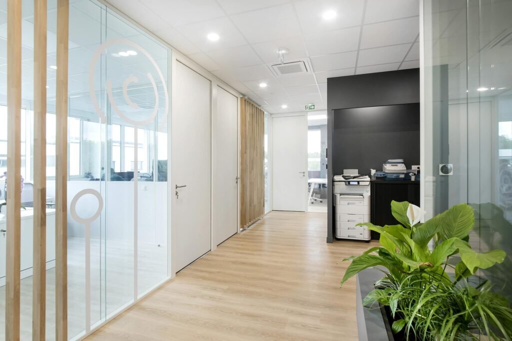 Couloir de bureau moderne avec des murs en verre, des sols en bois, une station d'impression et des plantes vertes près de l'entrée. La lumière naturelle pénètre par les grandes fenêtres à l'arrière-plan.