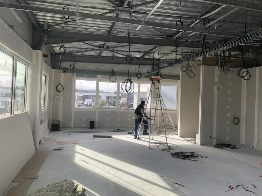 Une personne se tient sur une échelle et travaille à l'intérieur d'une pièce en construction, avec des câbles exposés, des murs inachevés et des matériaux de construction éparpillés sur le sol.