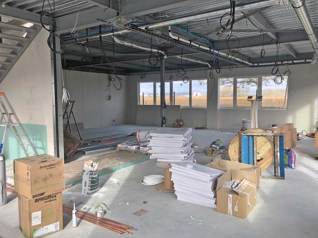 Intérieur d'un chantier de construction avec l'infrastructure exposée du plafond, les gaines, les câbles et les matériaux de construction empilés.