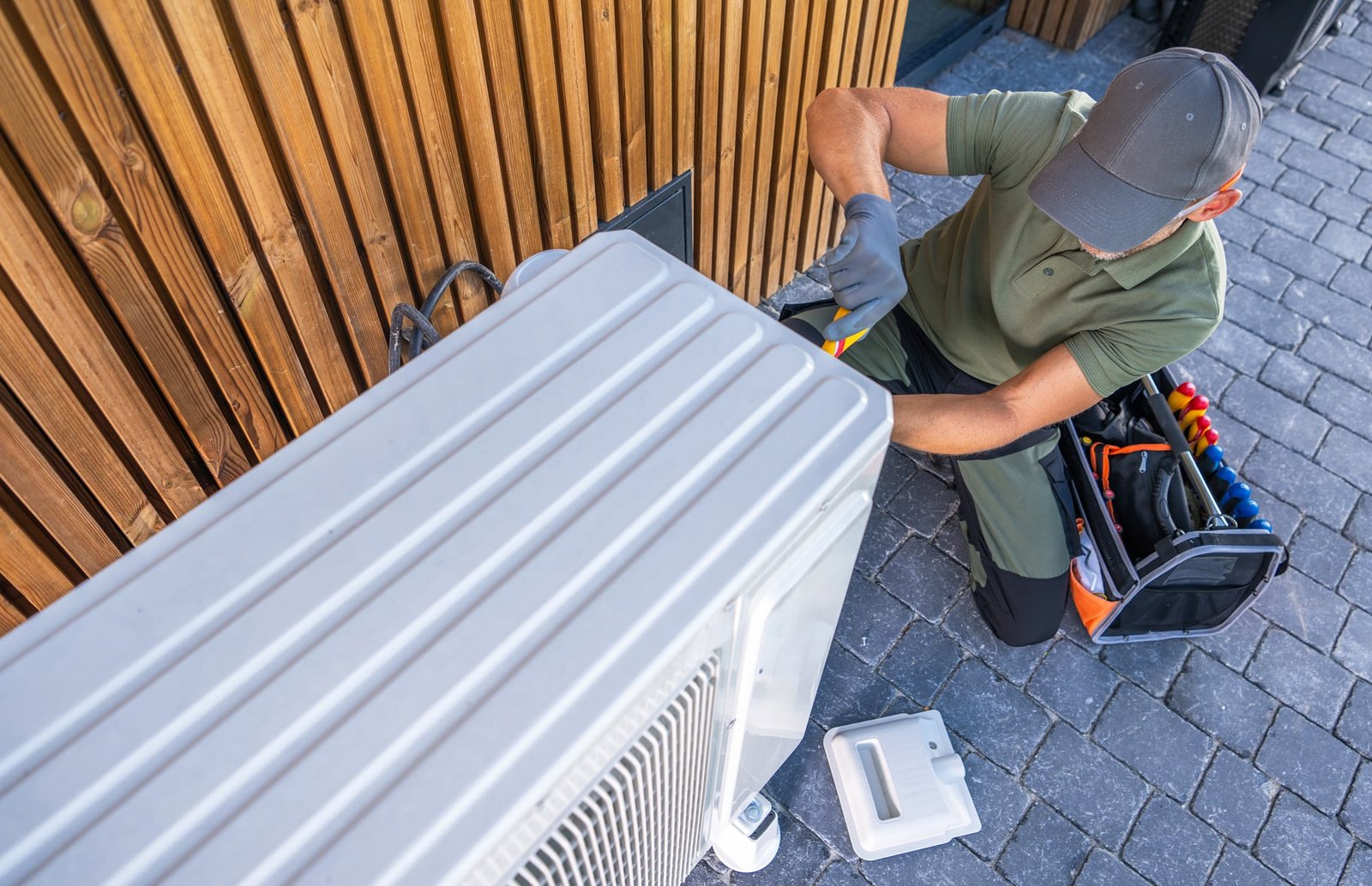 Un technicien portant des gants et une casquette répare une unité de climatisation extérieure près d'un mur en bois, avec un sac à outils et un panneau ouvert à proximité.