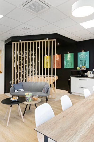 Salle de pause d'un bureau moderne avec des tables, des chaises, un coin salon, une kitchenette, des affiches colorées sur un mur noir, et des plantes sur un sol en bois clair sous des plafonniers lumineux.