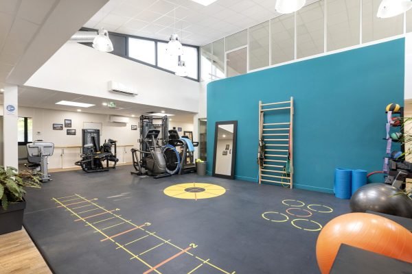 Salle de sport spacieuse avec des machines d'exercice, des barres murales, des miroirs, des ballons d'exercice et des tapis de fitness sur un sol noir avec des marquages, dans une pièce moderne et lumineuse.