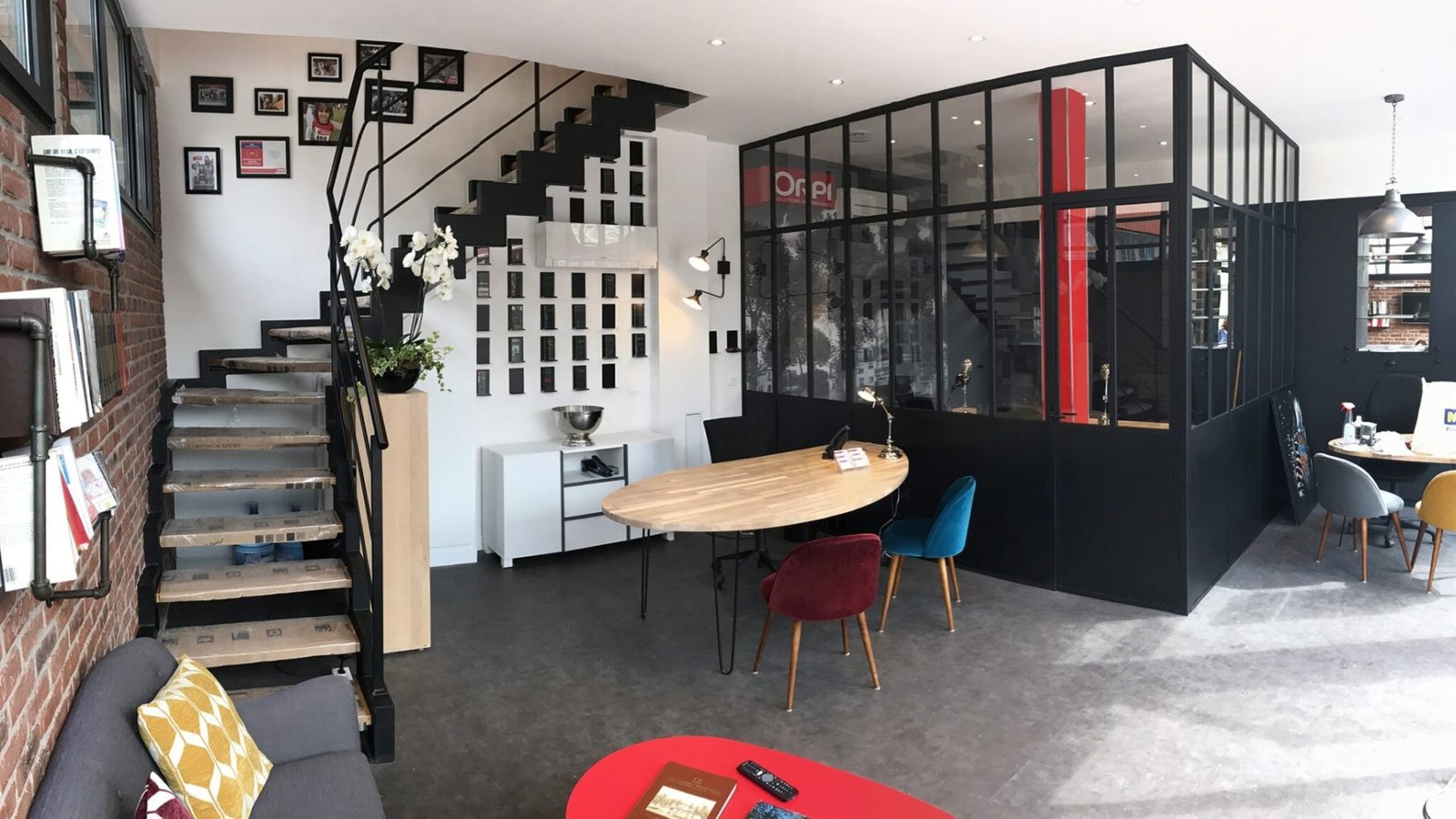 Espace de bureau moderne avec une salle de réunion vitrée, des escaliers avec des photos murales, une table ronde en bois avec des chaises colorées et divers objets de décoration.