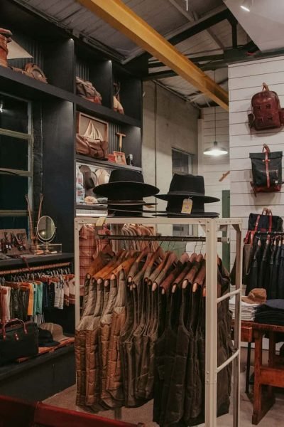Intérieur d'un magasin de vêtements avec des vestes, des chemises, des chapeaux et des sacs à dos soigneusement présentés sur des étagères, des rayonnages et des murs dans un cadre rustique et industriel.